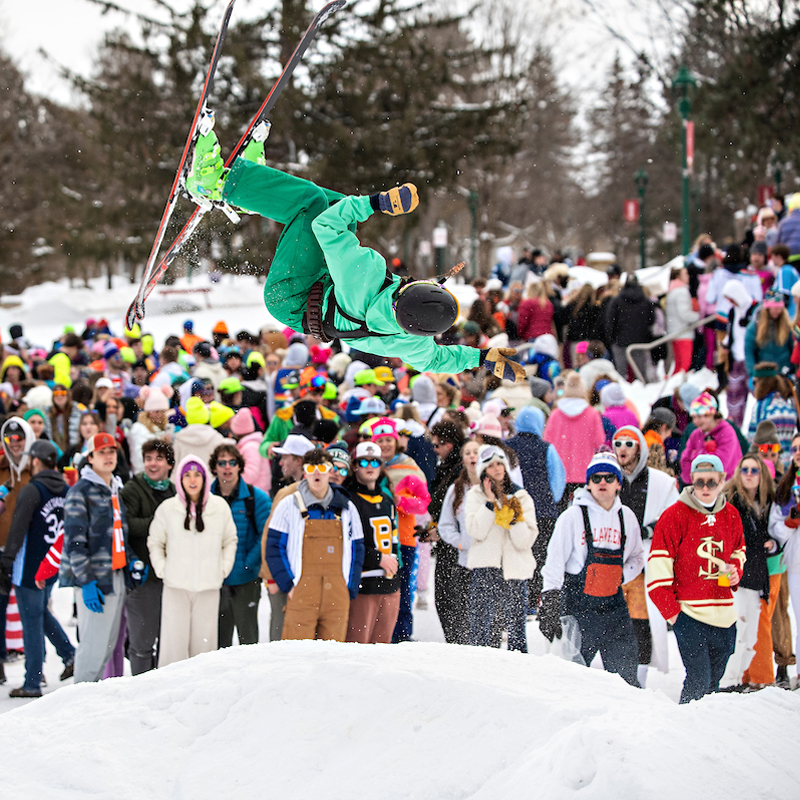 A student, wearing neon green snow gear and skis, sails through the air upside down on a snowy day as a full crowd of students look on in amazement.