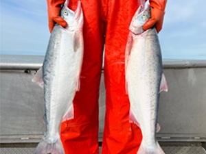 Jessica Normandeau, wearing bright red rubber waders, holds two large silver salmon while standing on a boat deck.