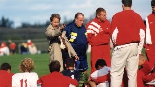 Bob Durocher, in a tan trench coat, is giving instructions to his team during a break in a game. The coach is gesturing with his hand while the players, dressed in red and white uniforms, sit and listen attentively. 