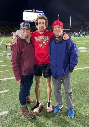 A young male athlete in a red St. Lawrence University t-shirt and black shorts stands on a football field at night, smiling with his arms around his parents. The woman is wearing a maroon jacket, jeans, and a colorful headband, while the man is dressed in a blue jacket and gray pants with a red beanie. The stadium lights and a scoreboard are visible in the background.